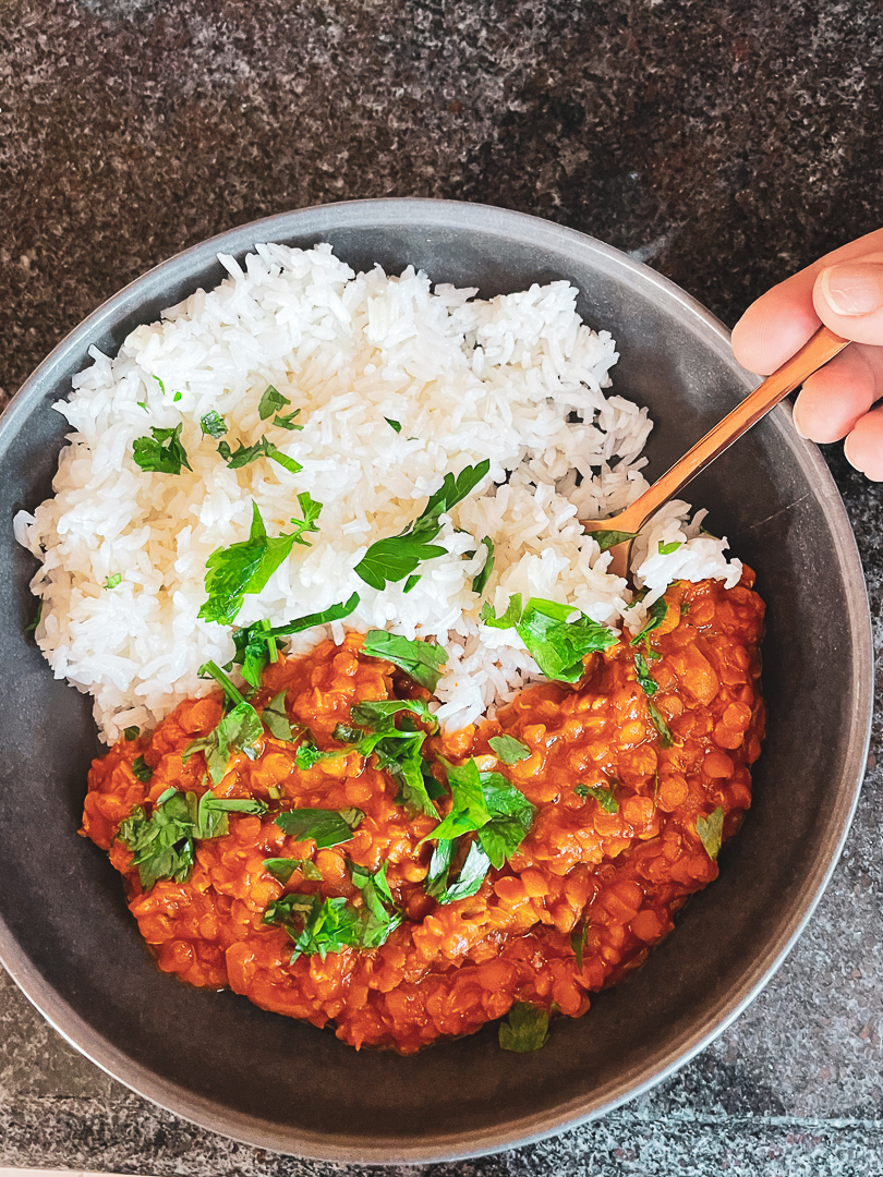Red Lentil curry in a bowl with white rice next to it garnished with parsley. Gold fork on side.